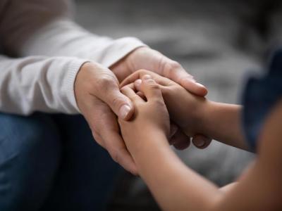 Close-up photo of a pair of adult hands cradling the folded hands of a child, as if to comfort them. Only hands and forearms are visible. 