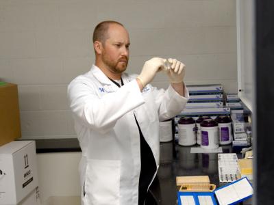 Kirby Mayer stands in a medical research lab; he's wearing a white lab coat and gloves and is holding up a microscope slide.
