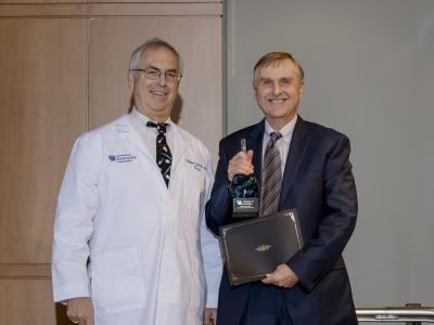 Dr. Philip Kern receives the 2023 Willard award. He's wearing a dark suit, holding a small statue and a certificate. Beside him is a man in a white medical coat. They're both smiling. 