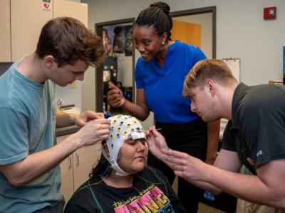 Dr. Lauren Whitehurst, a young Black woman with her hair in a bun, stands with two of her students who are setting up an electrode cap on the head of a research participant. 