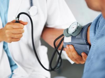 Image of someone in a white coat taking someone else's blood pressure. The photo is zoomed in on the blood pressure cuff and doctor's hands, so we can't see their faces. 