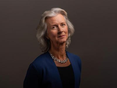 Portrait of an older white woman with wavy silver hair down to her shoulders; she's standing against a dark backgrop and giving slight smile. She's wearing a blue suit jacket, black scoop-neck shirt, and thick silver necklace. 