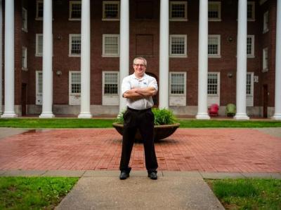 A middle-aged white man with glasses and short cropped brown and gray hair stands with his arm crossed, smiling at the camera, wearing black pants and a light gray polo shirt with white horizontal pinstripes and a UK logo embroidered on the upper left. Behind him we see a wide brick sidewalk, and beyond that a brick building with large white columns. 