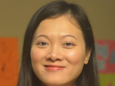 Headshot of a young asian woman with shoulder-length, straight black hair; she's smiling at the camera and wearing a dark coat jacket with light pink collared shirt. Behind her is a tan wall. 