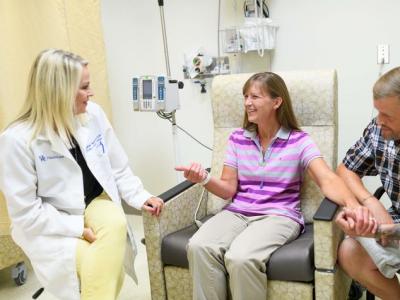 A blond research nurse in a white medical coat sits smiling beside a clinical trial participant who's in a treatment chair. The participant, a middle aged woman with brown hair, is smiling and holding the hand of her husband, who's sitting beside her. Behind her is infusion equipment (IV pole and pump) and medical equipment.