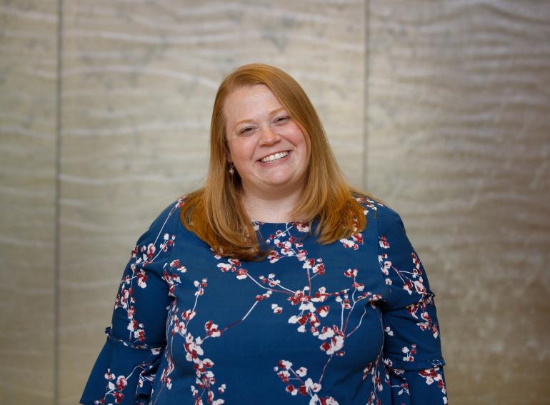 Allison Gibson, PhD, a white woman in her 30s, stands in front of a light gray wall, smiling at the camera. She has strawberry blond, straight hair past her shoulders, and she's wearing a long-sleeved blue dress with cherry blossoms. 