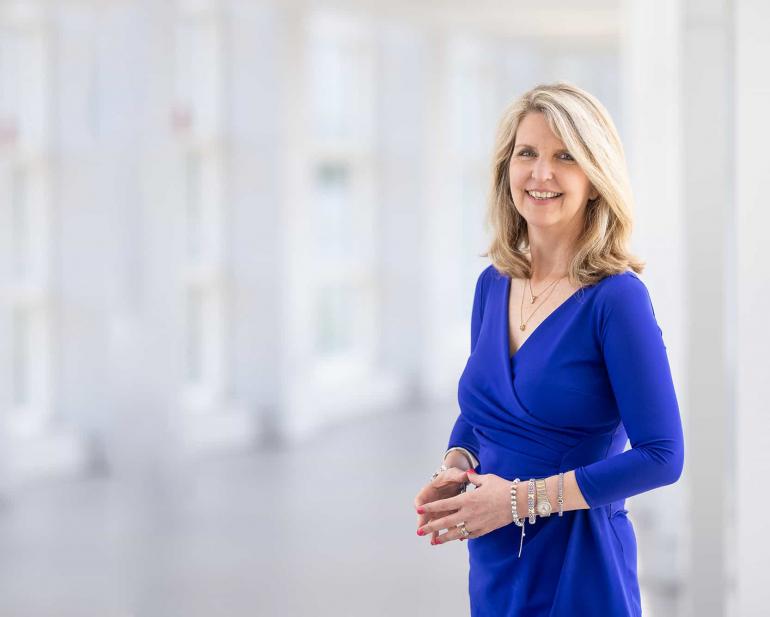 Dr. Susan Smyth, a white woman with blond, shoulder length hair, stands in a  white room with floor to ceiling windows behind her. She's wearing a royal blue v-neck dress and several bracelets. 