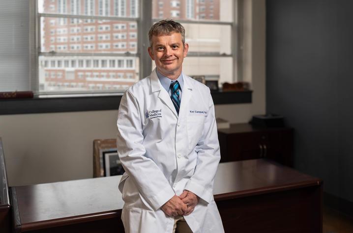 Ken Campbell, a middle-aged white man in a white lab coat, leans against a wooden desk in an office. He has light brown and gray hair and is smiling at the camera. He's wearing a light blue collared shirt with a navy and bright blue tie. There's a large window behind him, through which a big brick building is visible. 