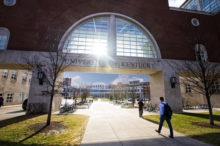 A brick and glass pedestrian bridge with a large half-moon window, under which are the words University of Kentucky. Light is shining through the window, creating a small sun burst. Two individuals are walking under the pedestrian bridge. 