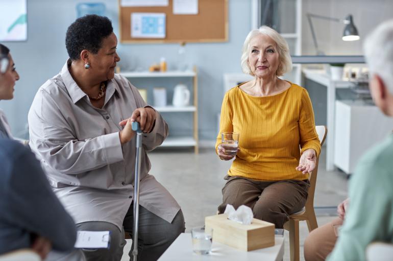 Two older women, one Black and one white, sit beside one another in a discussion circle in an office. The Black woman has short cropped hair and is holding her cane. The white woman has chin-length white hair and is holding a glass of water. We see the shoulders and hands of other members of the group discussion who are out of frame. 