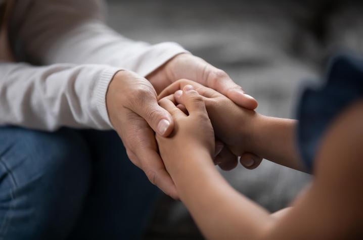 Close-up photo of a pair of adult hands cradling the folded hands of a child, as if to comfort them. Only hands and forearms are visible. 