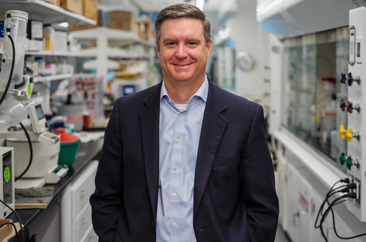 A middle-aged white man wearing a navy suit jacket and light gray collared shirt smiles at the camera; he's standing in a science lab. 