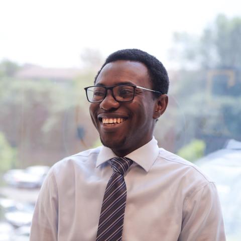 A young Black man is giving a big smile and looking slightly off camera. He has short hair, black rimmed glasses, and is wearing a cream colored collared shirt with a burgundy tie. He's standing in front of a window, with foliage visible behind him. 