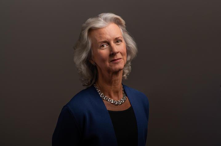 Portrait of an older white woman with wavy silver hair down to her shoulders; she's standing against a dark backgrop and giving slight smile. She's wearing a blue suit jacket, black scoop-neck shirt, and thick silver necklace. 