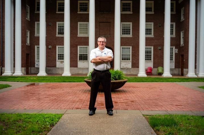 A middle-aged white man with glasses and short cropped brown and gray hair stands with his arm crossed, smiling at the camera, wearing black pants and a light gray polo shirt with white horizontal pinstripes and a UK logo embroidered on the upper left. Behind him we see a wide brick sidewalk, and beyond that a brick building with large white columns. 