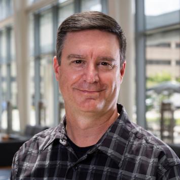 Headshot of Tom Prisinzano, smiling with a wall of windows blurred into the background. He's a white man, middle-aged, with short, dark brown hair and brown eyes. He's wearing a plaid shirt, deep purple and gray, buttoned almost to the top with a black shirt beneath. 