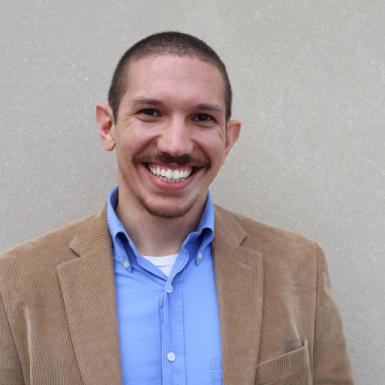 Photo of Anthony Mangino, a young man with buzzed, dark hair and a dark mustache. He's wearing a beige suit jacket with a light blue collared shirt underneath. He's smiling at the camera an standing in front of a light gray wall. 