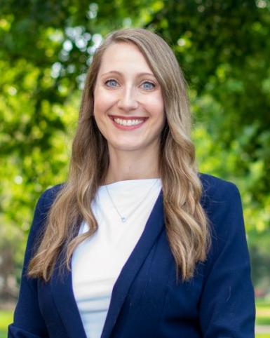 Headshot of Rosie Shrout, a young white woman with long blond hair. She's wearing a dark blue blazer with a white shirt. There are lush trees blurred in the background. 