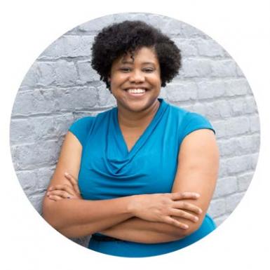 photo of Lea Whitehurst-Gibson, a young Black woman with short curly hair and a radiant smile. She's wearing a light blue, short-sleeved shirt with a drapey neckline; she's leaning against a gray brick wall and her arms are folded across her chest. 