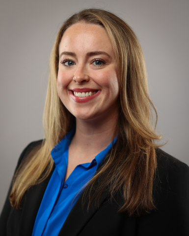 Professional headshot of a white woman in her 30s, with long, straight dark blond hair; she's smiling at the camera, wearing read lipstick, a black suit jacket, and a blue shirt. 