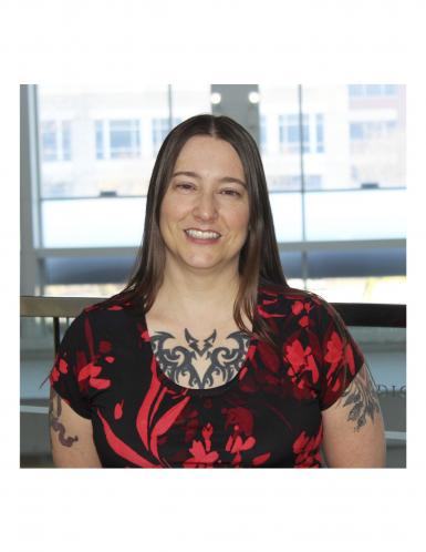 Photo of a white woman with long, straight brown hair. She's smiling at the camera, wearing a black and red floral scoop-neck shirt, and has tattoos on her chest and arms. 