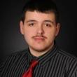 Professional headshot of a young white man with dark, short hair, dark brown eyes, and a thin dark mustache. He's wearing a gray and black striped shirt with a red satin tie. 