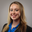 Professional headshot of a white woman in her 30s, with long, straight dark blond hair; she's smiling at the camera, wearing read lipstick, a black suit jacket, and a blue shirt. 