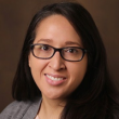 Professional headshot of a woman with long, straight, black hair and rectangular black-rimmed glasses. She's smiling, wearing a light pink blouse and gray cardigan, and behind her is a dark brown backdrop. 