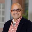 Headshot of a middle-aged man, bald with thick-rimmed rectangular glasses, smiling at the camera wearing a pink floral shirt and dark suit jacket. 