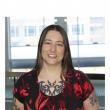 Photo of a white woman with long, straight brown hair. She's smiling at the camera, wearing a black and red floral scoop-neck shirt, and has tattoos on her chest and arms. 